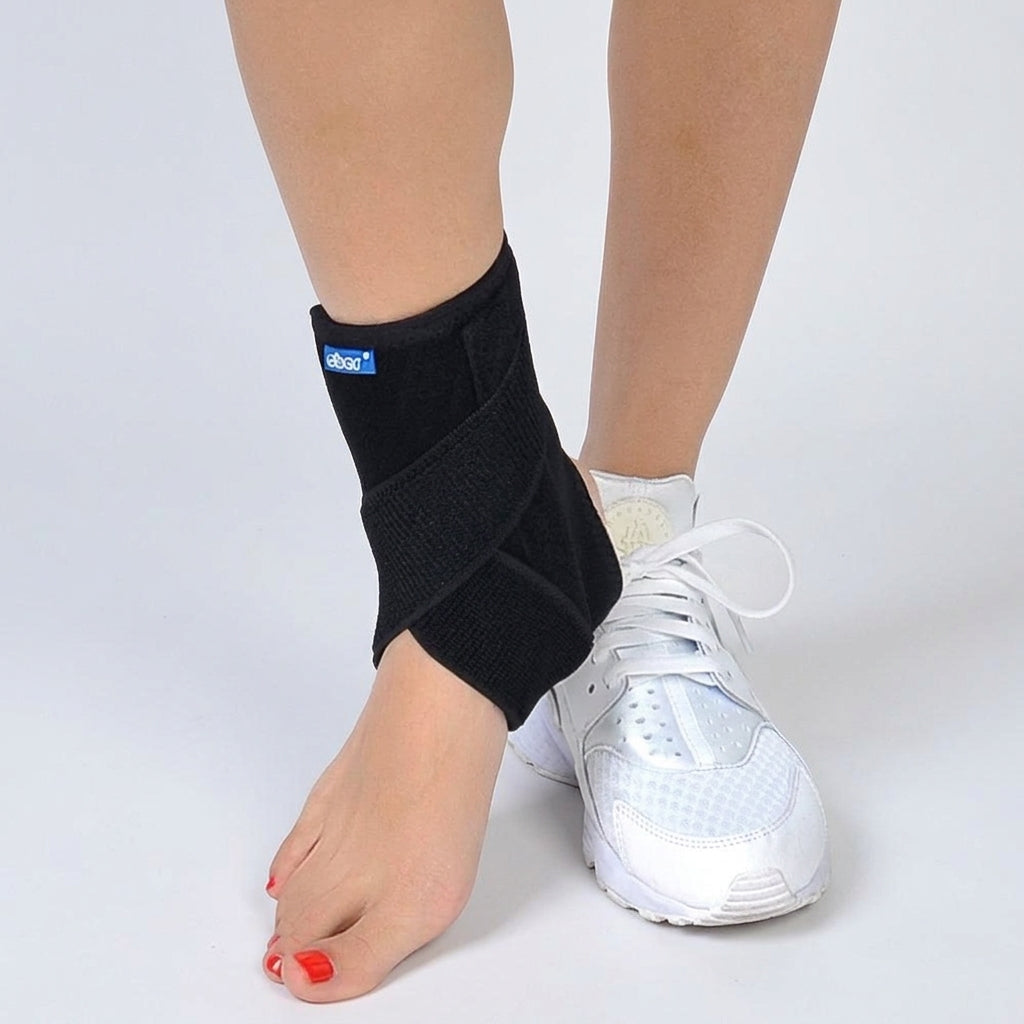 A ankle brace torn ligament worn on a person's left ankle, shown with bare foot and white athletic shoe on a light gray studio background.
