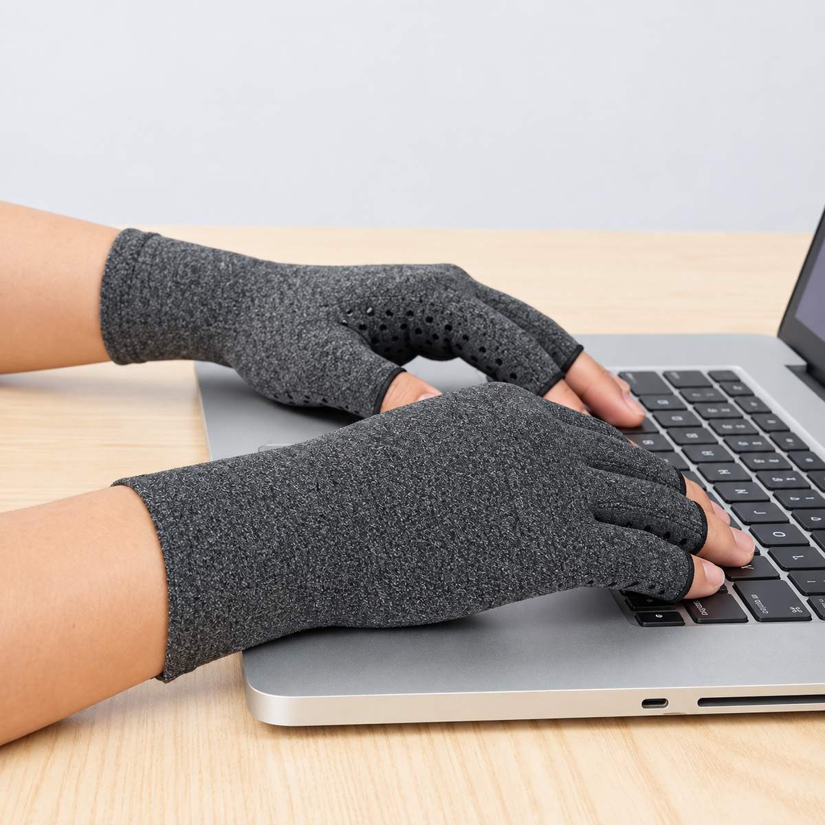 A person wearing hand joint pain compression gloves while typing on a silver laptop at a light wood desk.