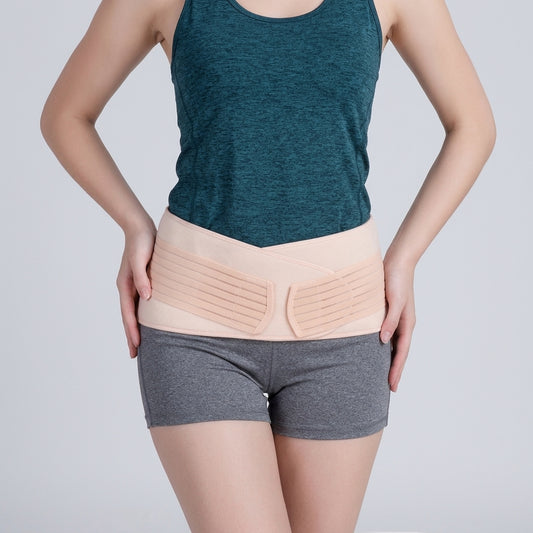 A woman wearing postpartum pelvic recovery belt over a dark teal tank top and gray athletic shorts, shown from the chest down against a light gray studio background.