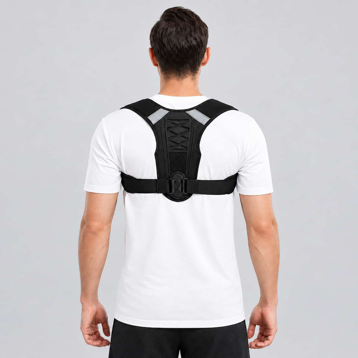 A man viewed from behind wearing an upper back support brace with reflective gray shoulder straps over a white t-shirt, set against a solid gray studio background.